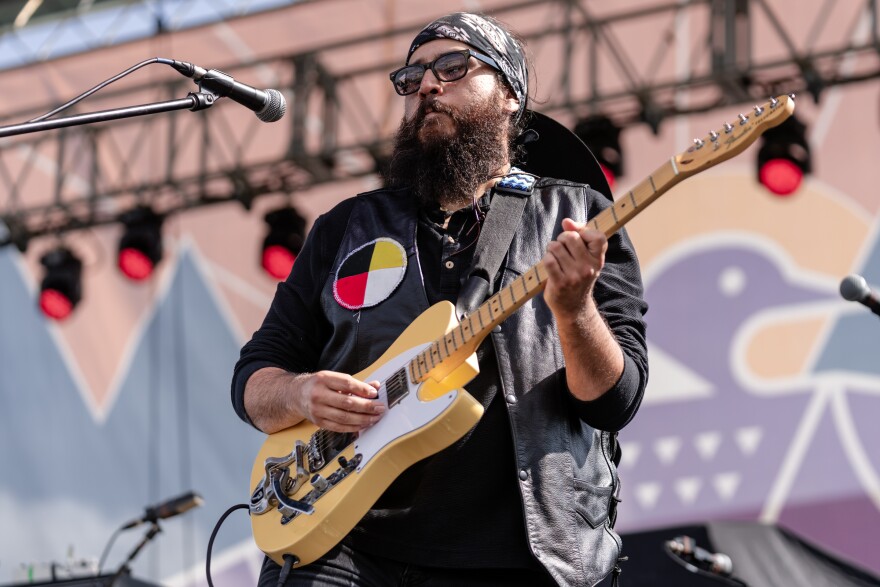 A man dressed in black playing a yellow guitar. He is on stage with a large purple loon backdrop. 
