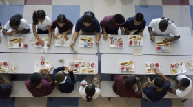 Una toma de cámara desde arriba muestra las comidas escolares frente a unos 15 niños sentados en una mesa larga.
