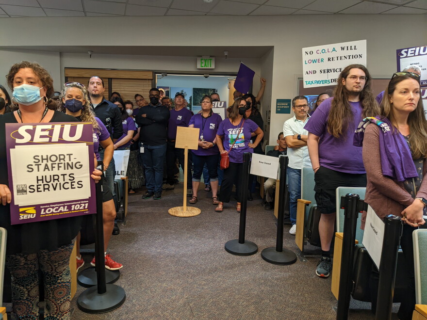 People wearing purple shirts and holding signs demanding better pay and more staffing fill the chambers of a local government chambers.