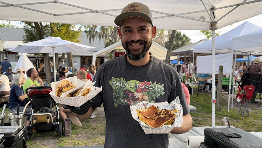 Ivan Perez, owner and chef of Taino Roots Artisan Kitchen, happily serves guests on Feb. 23 at the GNV Market at Heartwood. He is posed with an order of braised chicken tacos (left) and braised chicken quesadillas (right). (Miguel Molina/WUFT News)