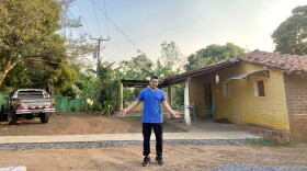 A man in a blue shirt stands in front of a house on a dirt road with his arms out