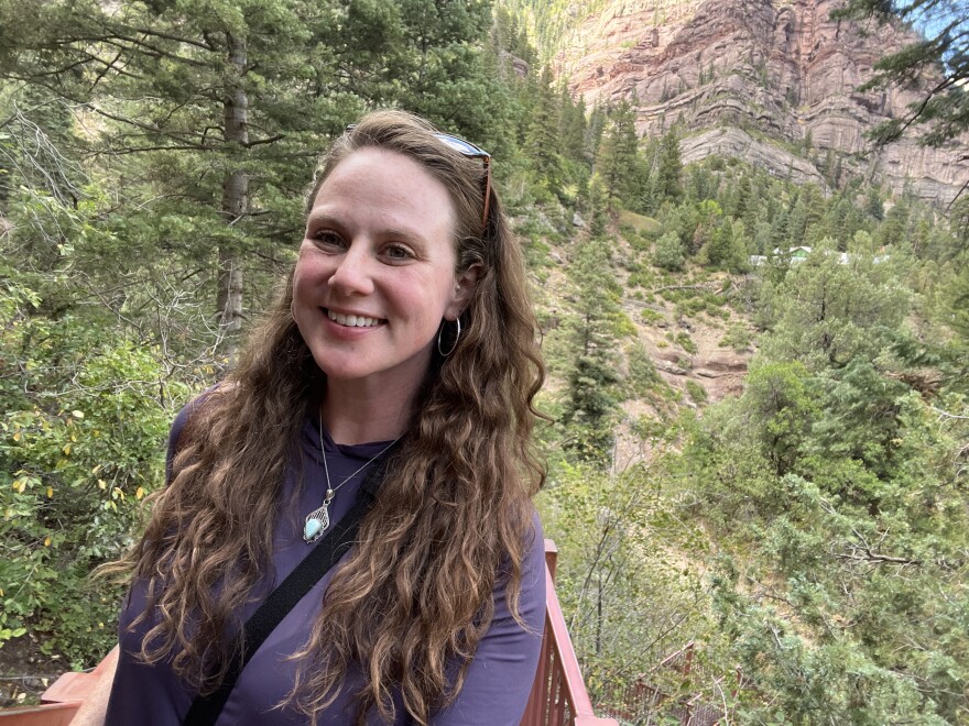 A woman with long hair smiles at the camera as she stand out in the wilderness.