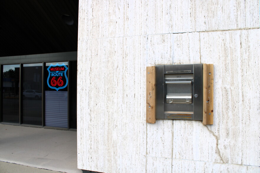 A metal mail slot is mounted on a white stone wall. To the left, a glass door has a neon Route 66 Museum sign. The area appears quiet and unoccupied.