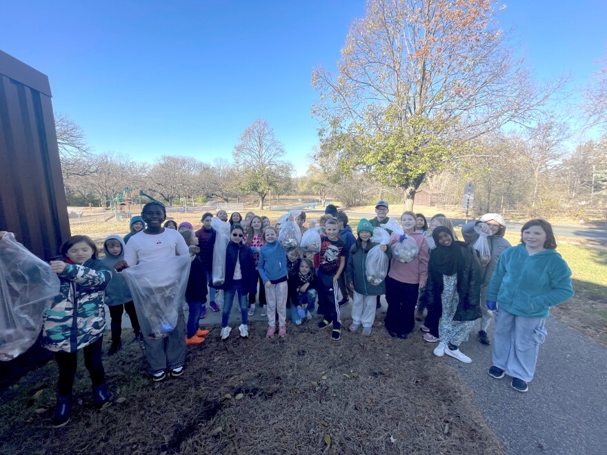A large group of students stands outside of their school, holding clear plastic bags filled with trash.