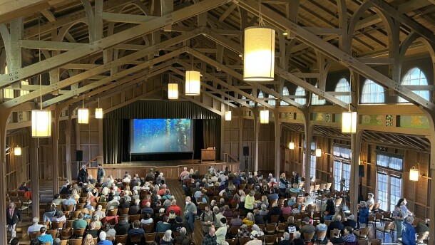 A crowd of people gather in a wooden hall adorned with hanging lights and many windows. 