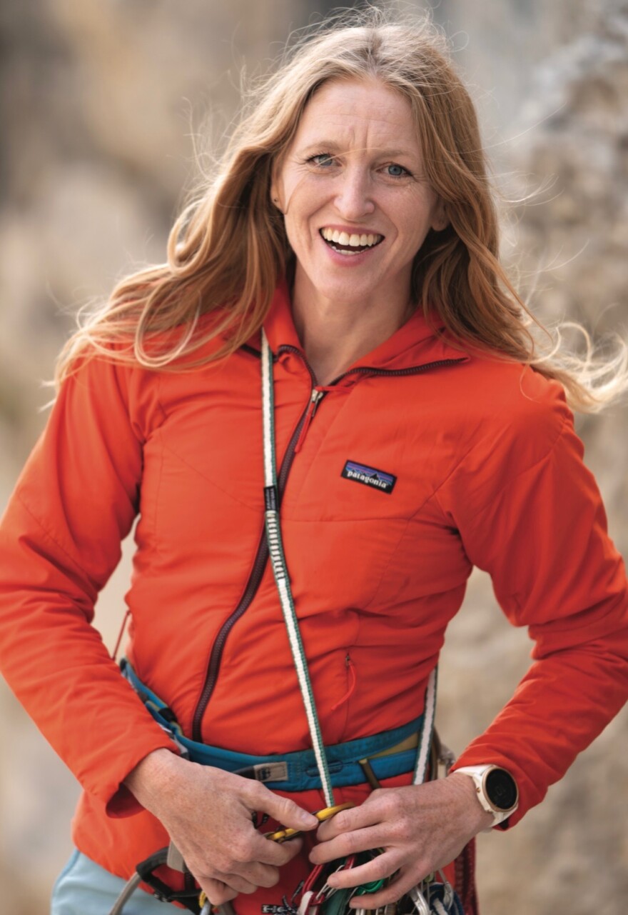 A woman with long red hair smiles at the camera, wearing a climbing harness and a sling with technical climbing year.