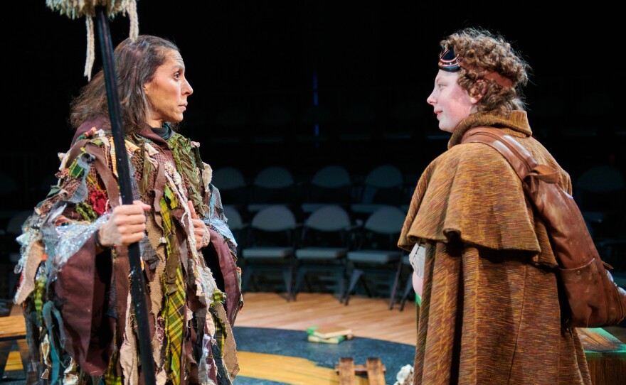 An actor dressed in rags and holding a staff looks intently at a younger actor wearing goggles on top of his head and a leather satchel.