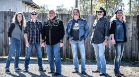 Group of older white men wearing jeans and flannel or button-down shirts standing outside in front of a fence 