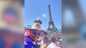 Jeff Toy is pictured in the crowd at the Olympic Games, he is in the foreground with the Eiffel Tower in the background. Toy wears a stars and stripes T-shirt and a straw sunhat. Perched on top of the sunhat is a stuffed bald-eagle. Toy is looking at the camera and wears sunglasses.