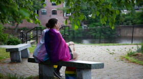 Two people wearing Pride flags around their shoulders sit on an outdoor bench during a Pride Month event.