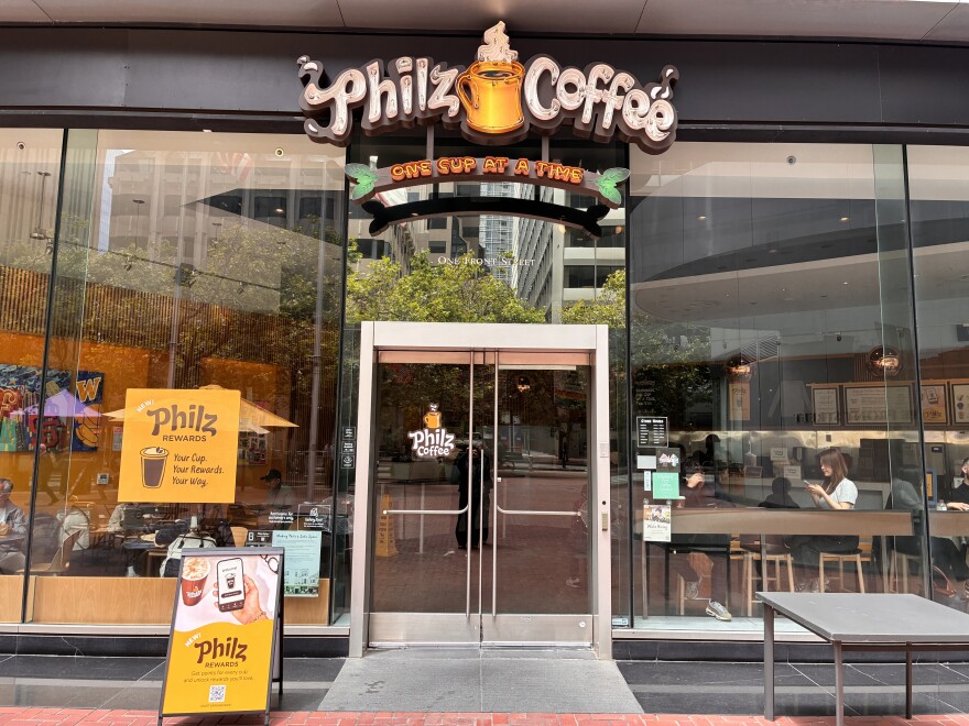 Photo of the outside of a Philz Coffee in San Francisco. The company's logo is above two glass doors, underneath that, an orange neon sign reads: "One cup at a time." The store has large glass doors and walls so you can see people sitting down inside. 