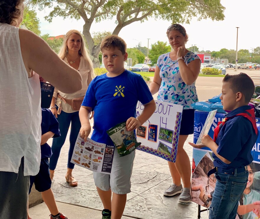 Young boys sell popcorn at a table outside with women standing behind them.