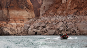 A small boat skims across the blue water of a lake. Behind the boat are large cliffs with white markings up to the middle of the cliff. Above the white markings, the cliffs are orange.