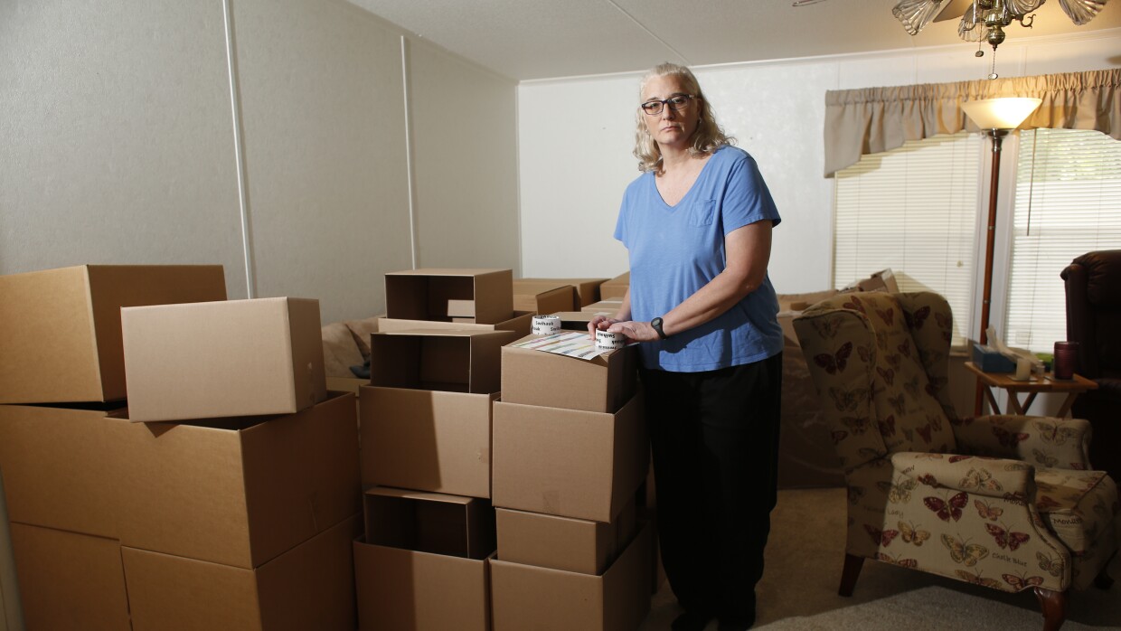 A woman with white hair wearing a blue shirt stands besides boxes in her living room. 