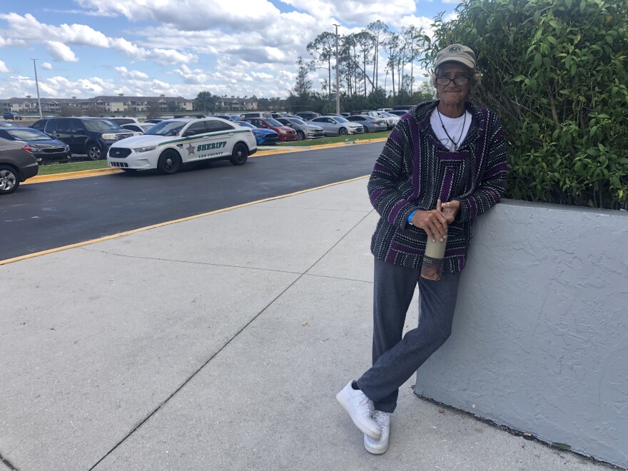 Gary Scot Janikula standing on pavement leaning on a cement wall up that comes up to his waist that contains some green plants surpassing his head. He's wearing white shoes, blue pants and a dark multi-colored sweater and a beige cap. Behind him is the Hertz arena parking lot with a Lee County Sherriff's SUV. The sky behind him is blue with many white fluffy clouds.