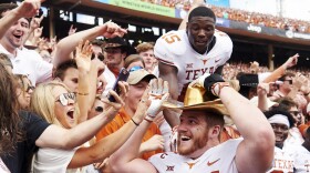 A smiling football player in a white jersey with orange numbers wears a golden hat in front of another player and a crowd of fans in a stadium.