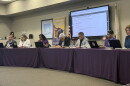 District 87 school board members and the superintendent sit at a long table draped in purple with laptops and nameplates, beneath a screen showing the Oct. 21 meeting agenda and beside a Bloomington Public Schools banner.