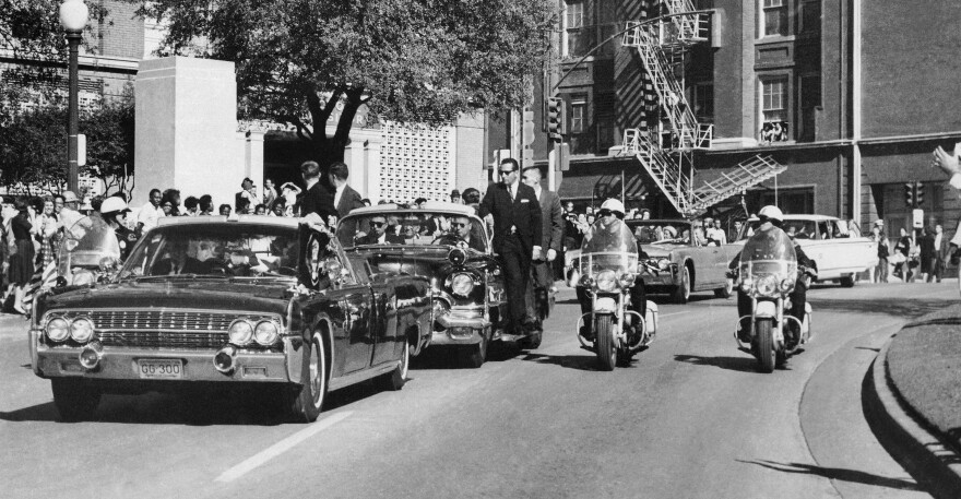 In this Nov. 22, 1963, file photo, seen through the foreground convertible's windshield, President John F. Kennedy's hand reaches toward his head within seconds of being fatally shot as first lady Jacqueline Kennedy holds his forearm as the motorcade proceeds along Elm Street past the Texas School Book Depository in Dallas. The license plates on the vehicle, which were discarded when the vehicle was sent for upgrades, are going up for auction. 