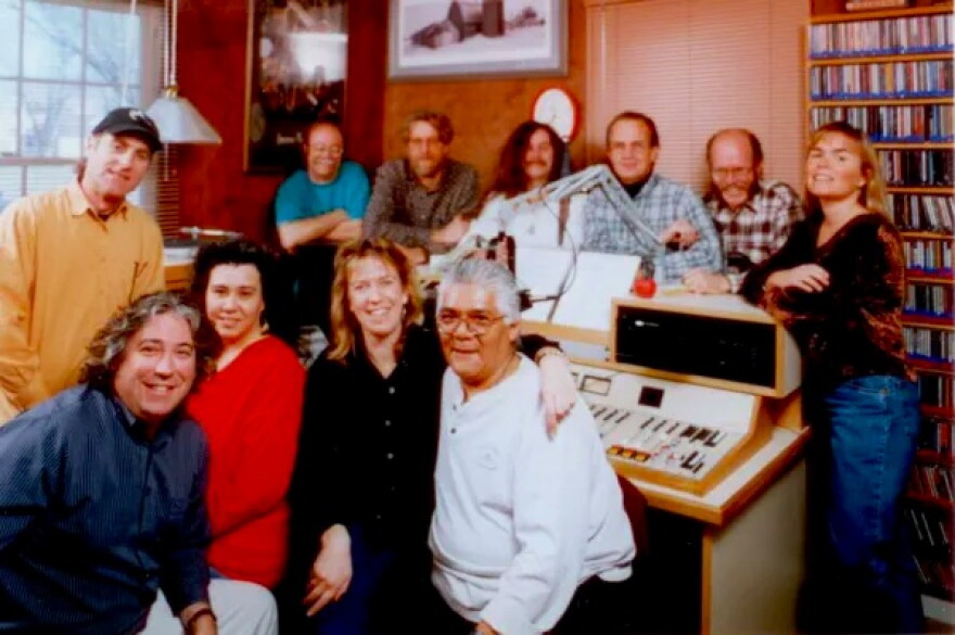 Three smiling people face the camera. The person in the center has their arms around the shoulders of the other two.