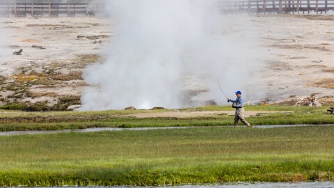 A man walks across a river landscape casting a fishing rod into the water as a geyser explodes behind him.