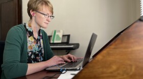 A woman sits at a desk typing on her computer and has earphones plugged in