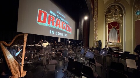 A photo taken from the wings of the Missouri Theatre stage, showing the empty chair setup for 80 musicians. A harp is featured prominently. A large projector screen with the opening of Dreamworks' 'How to Train Your Dragon' is above the percussion section.