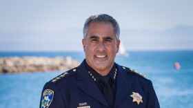 A man in a formal blue police uniform stands with his hands clasped in front of him. He's in front of a railing and behind that is the ocean with a jetty in the distance.