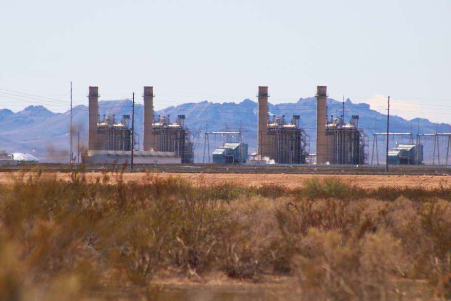 A power plant with several buildings, photographed form across a dry field. There are tall mountains in the background.