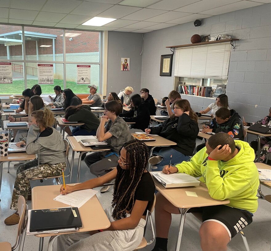 High school students are sitting at their desks in a classroom. Around 20 students can be seen in a gray room with a large window in the far side of the classroom.