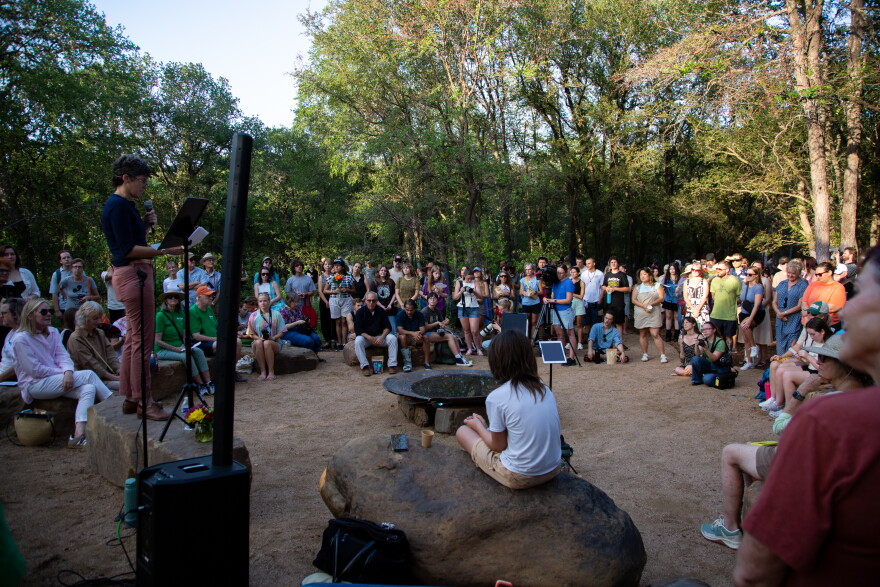 A person speaks on a small stage outside with a crowd sitting on the ground and standing watching.