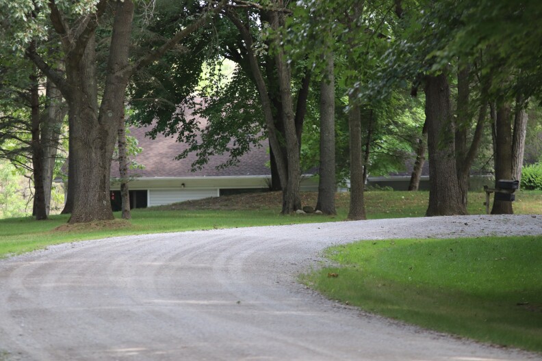 In the distance, a gray roof line of a pale yellow house is mostly blocked from view by thick forests. A gravel driveway curves in front. 