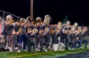 High school students play brass instruments on a football field. They are arranged in two rows. The front row kneels on one foot and the back row stands behind them.