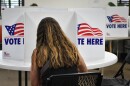 Interior photograph of a person sitting inside a room. They are seated in a folding chair at round table where there are "Vote Here" signs. They appear to be writing. 