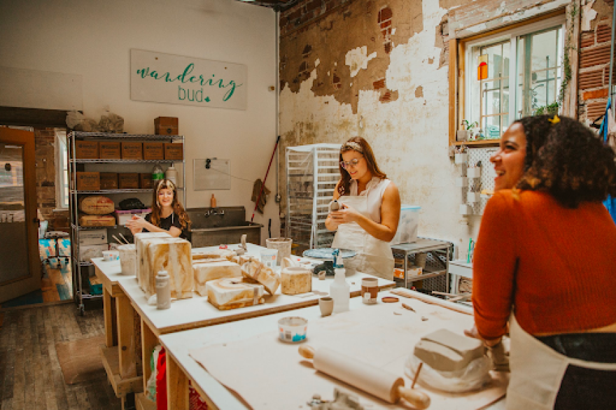 Three women wearing aprons smile while working around a large tables filled with ceramic making supplies.