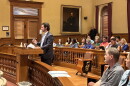 A man in a suit speaks at a podium in a courtroom filled with seated people. A large portrait hangs on the wall behind him. Papers are on the podium and benches.