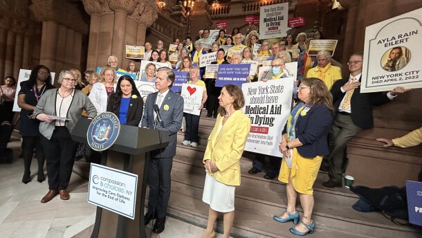 State Sen. Brad Hoylman-Sigal, at lectern, and Assemblymember Amy Paulin, in yellow coat, the sponsors the Medical Aid in Dying Act, are surrounded by lawmakers and advocates on Monday, June 9, 2025, at the New York State Capitol ahead of the Senate's vote on the bill.