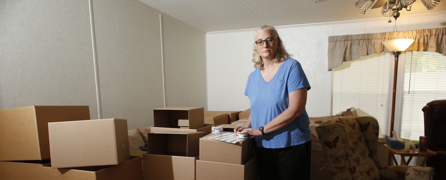 A woman with white hair wearing a blue shirt stands besides boxes in her living room. 