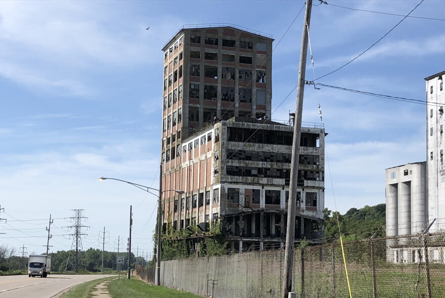An abandoned, multi-story industrial building with broken windows stands behind a chain-link fence next to a highway.