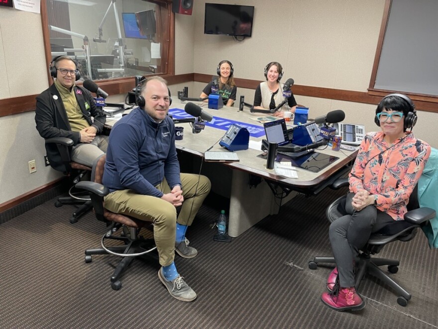 Five smiling people wearing headphones sit at a table in a radio talk studio: a man at left foreground has short brown hair and is wearing glasses, a black blazer with a hot pink bicycle pin on the lapel, an olive green sweater and beige pants; a man at right foreground has short brown hair and is wearing a blue fleece, tan pants, bright blue socks and grey shoes; a woman at background left has long brown hair and is wearing a black t-shirt with green lettering; a woman at background right has short brown hair and is wearing a beige vest over a black shirt; a woman at center has short dark hair and is wearing turquoise glasses, a pink patterned button-down shirt, black pants and hot pink boots.