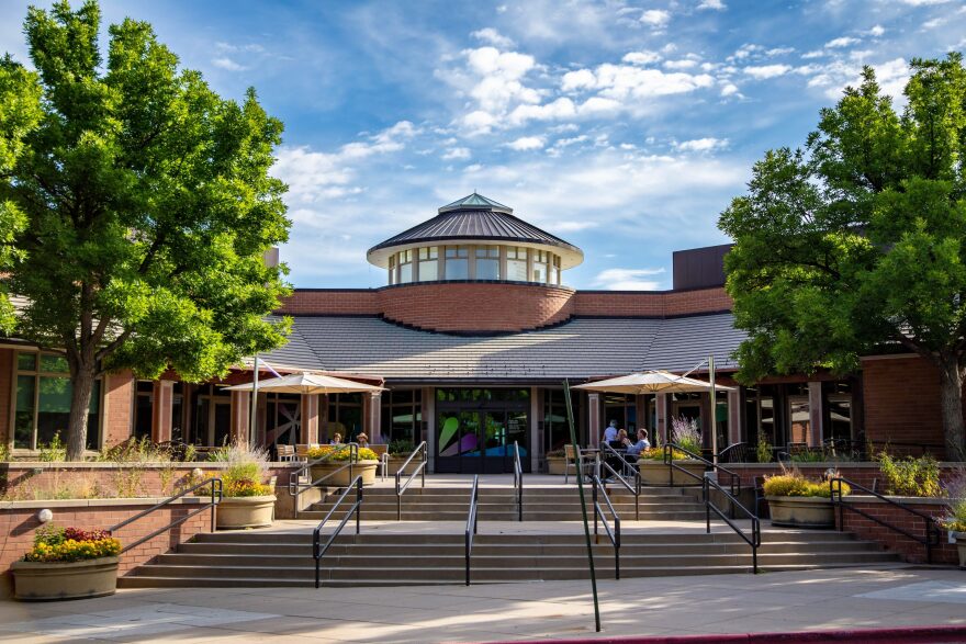 The outside of a brick building. Stairs lead to the doorway. Outside there are lots of flowers and green trees.