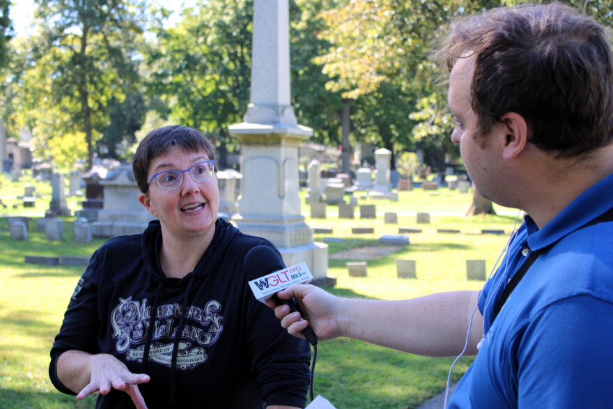 A man with a microphone interviews a woman in a cemetery. 