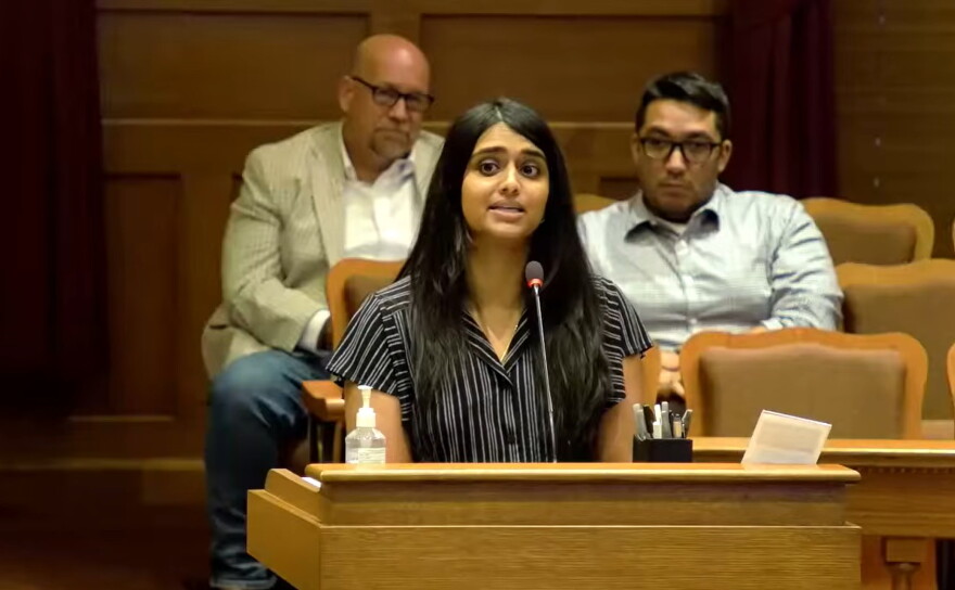 A woman speaks at a podium in a wood-paneled room, with two men sitting and listening in the background. A microphone, hand sanitizer, and papers are on the podium.