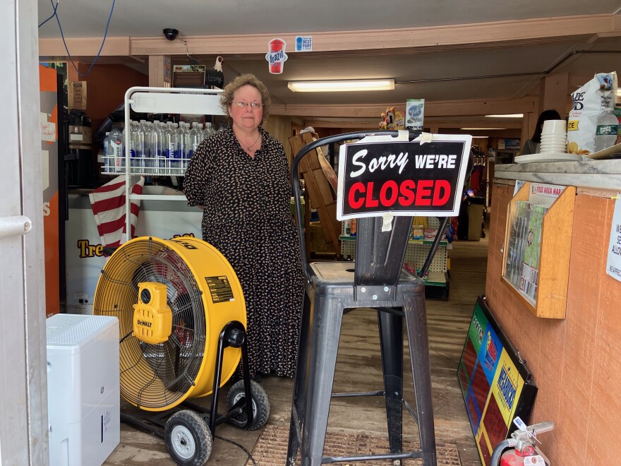 A woman standing inside a general store with a fan to her right and a 'Sorry we're closed' sign to her right.