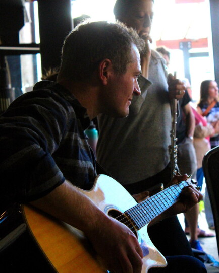 A man with his guitar in the foreground and a flute player behind him.