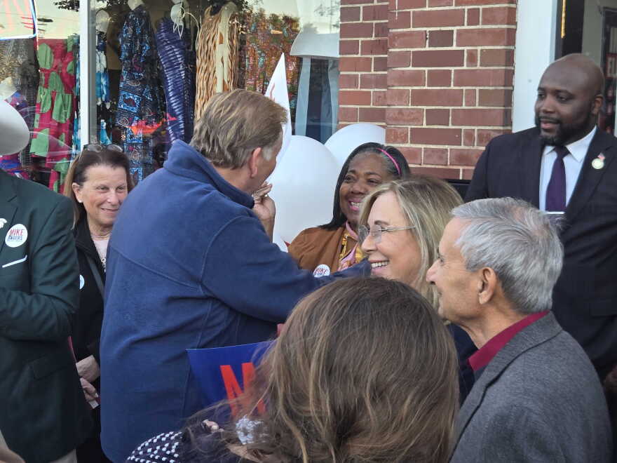 An outdoor campaign gathering where several people interact in front of a brick building. In the center, a man wearing a blue fleece jacket stands with his back partially to the camera, greeting and interacting with a smiling woman with blonde hair and glasses. Other smiling onlookers surround them. Some individuals in the crowd wear campaign buttons that read "Mike Rogers." In the background, a shop window displays colorful clothing, and white campaign balloons are partially visible.