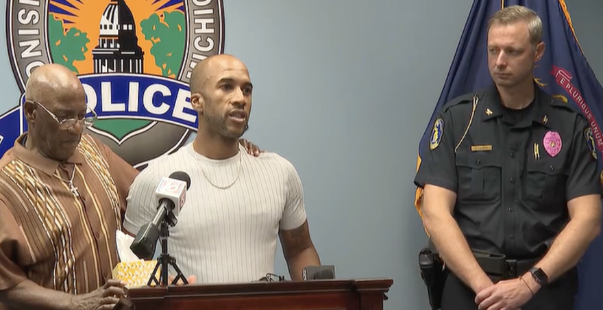 Two men stand at a podium and speak into a microphone next to a police officer