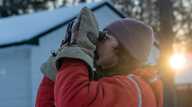 A man in a red coat and large mittens peers through binoculars on a winter morning.