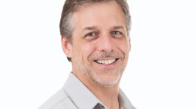 A smiling middle-aged man with short graying hair and a trimmed beard poses against a plain white background. He is wearing a light gray collared shirt and looking slightly to the side.