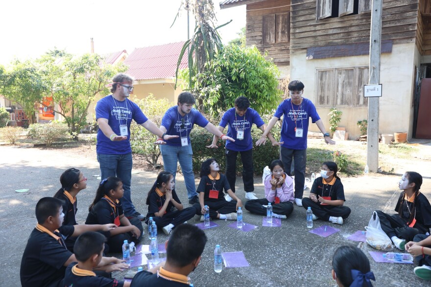 Four people in blue shirts and hanging lanyards stand over a group of students clad in black.