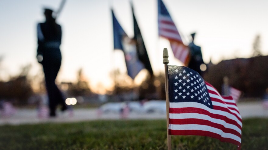 A small American flag pokes out of the ground, with other flags and two marching people in the background. 